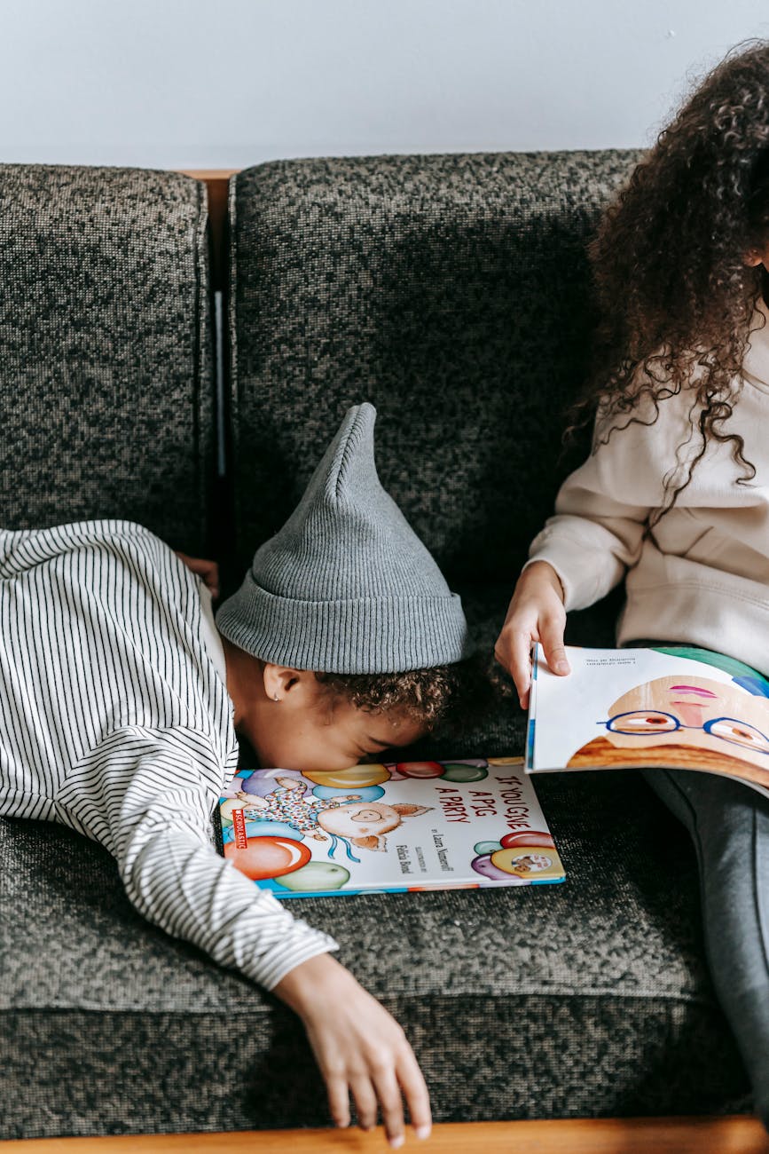 unrecognizable cute little ethnic siblings studying colorful books on sofa
